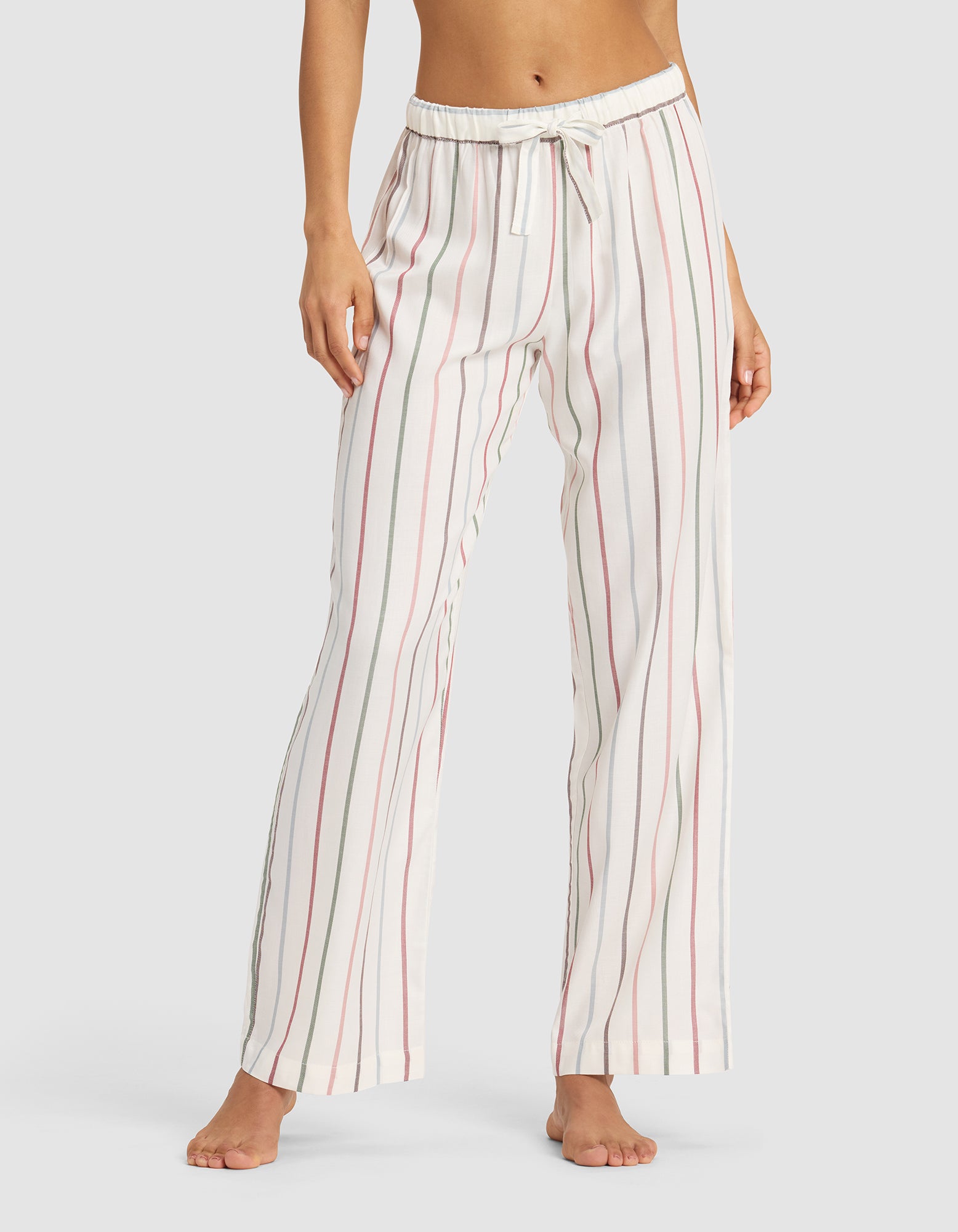 A woman models Cozy Earth’s Women's Soft Woven Pajama Pant—loose-fitting, white with vertical pink and gray stripes—barefoot against a simple backdrop.|Color:Cottage Stripe