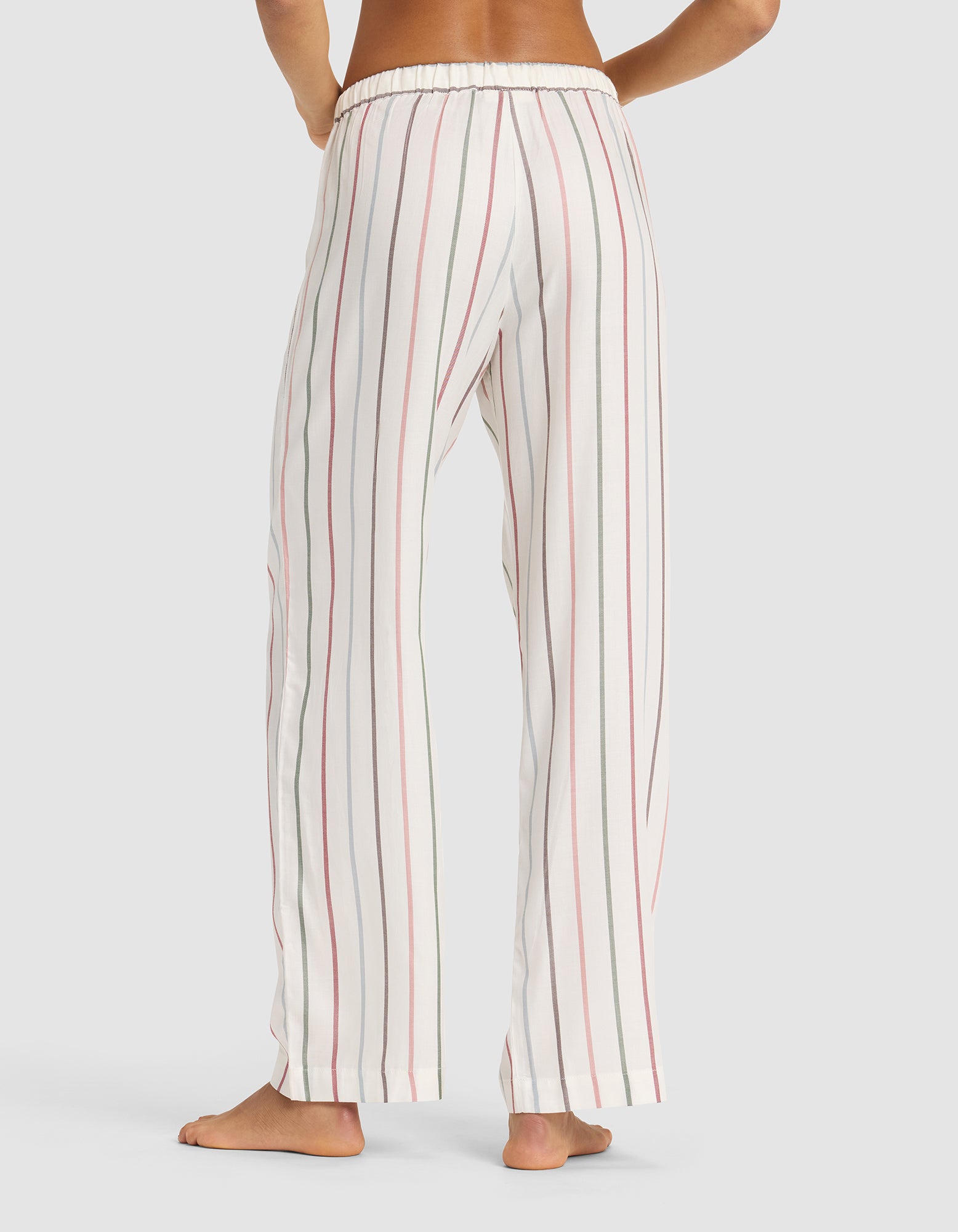 A person stands barefoot, shown from behind, wearing Cozy Earth Women's Soft Woven Pajama Pant—loose-fitting white pants with vertical red and black stripes—on a plain light background, displaying only the lower back and legs.|Color:Cottage Stripe