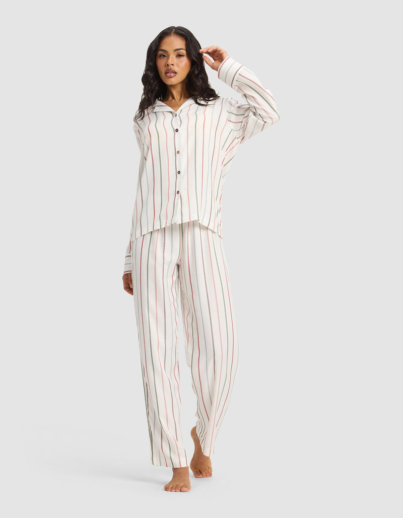 A woman stands barefoot against a plain background, wearing Cozy Earth's Women's Soft Woven Pajama Pant in a white fabric with vertical red and grey stripes. The pants are loose-fitting and paired with a matching button-up, collared pajama top.
