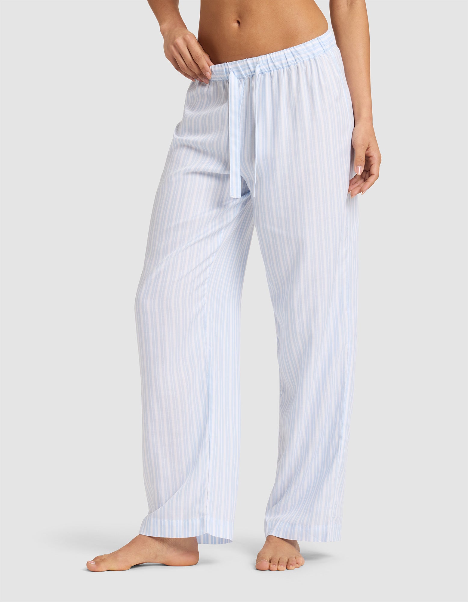 A woman stands barefoot in Cozy Earth Women's Soft Woven Pajama Pant, featuring light blue and white vertical stripes, an elastic waistband, and a drawstring, against a plain light gray background. 