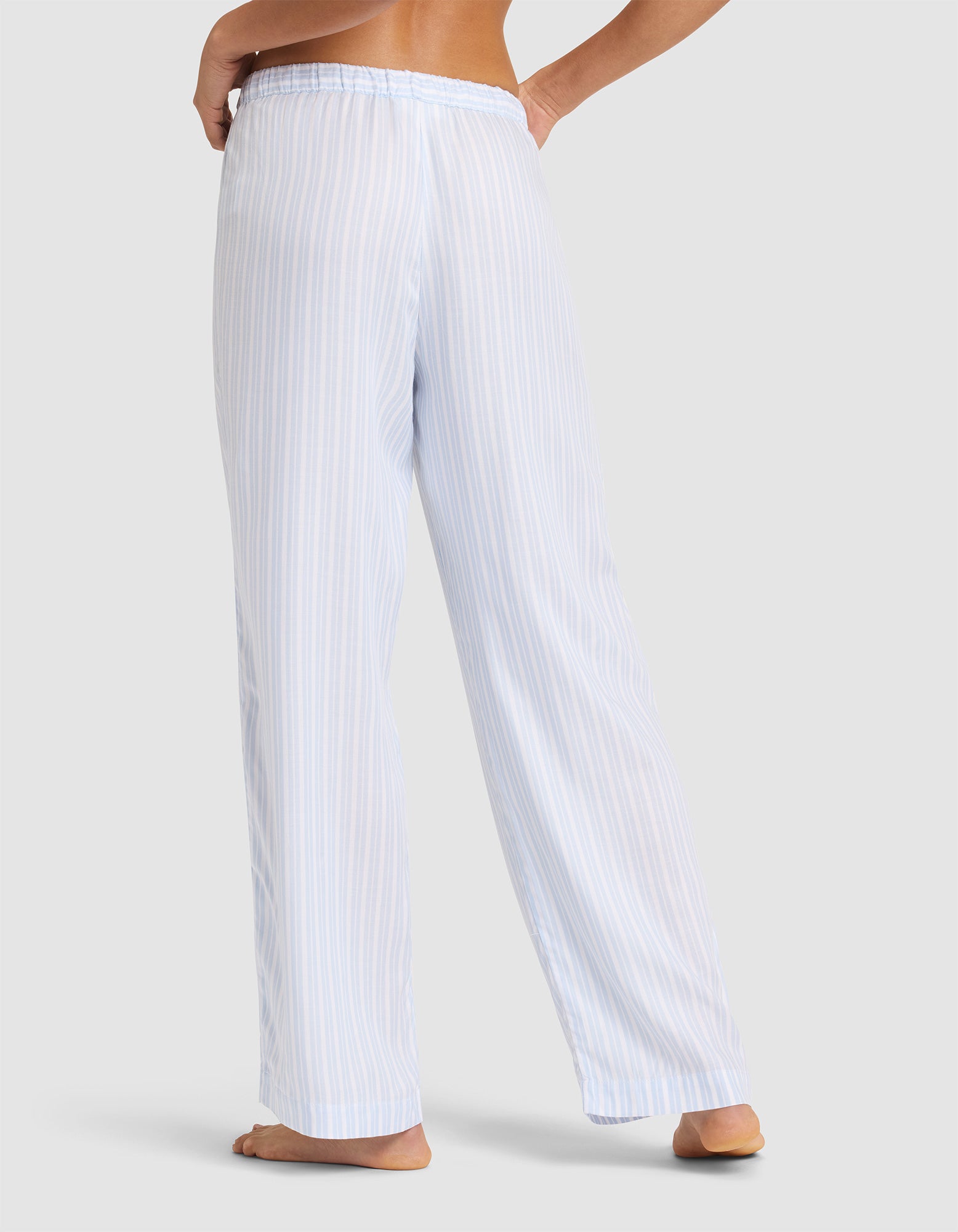 A woman models Cozy Earth’s Women’s Soft Woven Pajama Pant, featuring loose-fitting, light blue and white vertical stripes, shown barefoot from the waist down against a plain light gray background.  