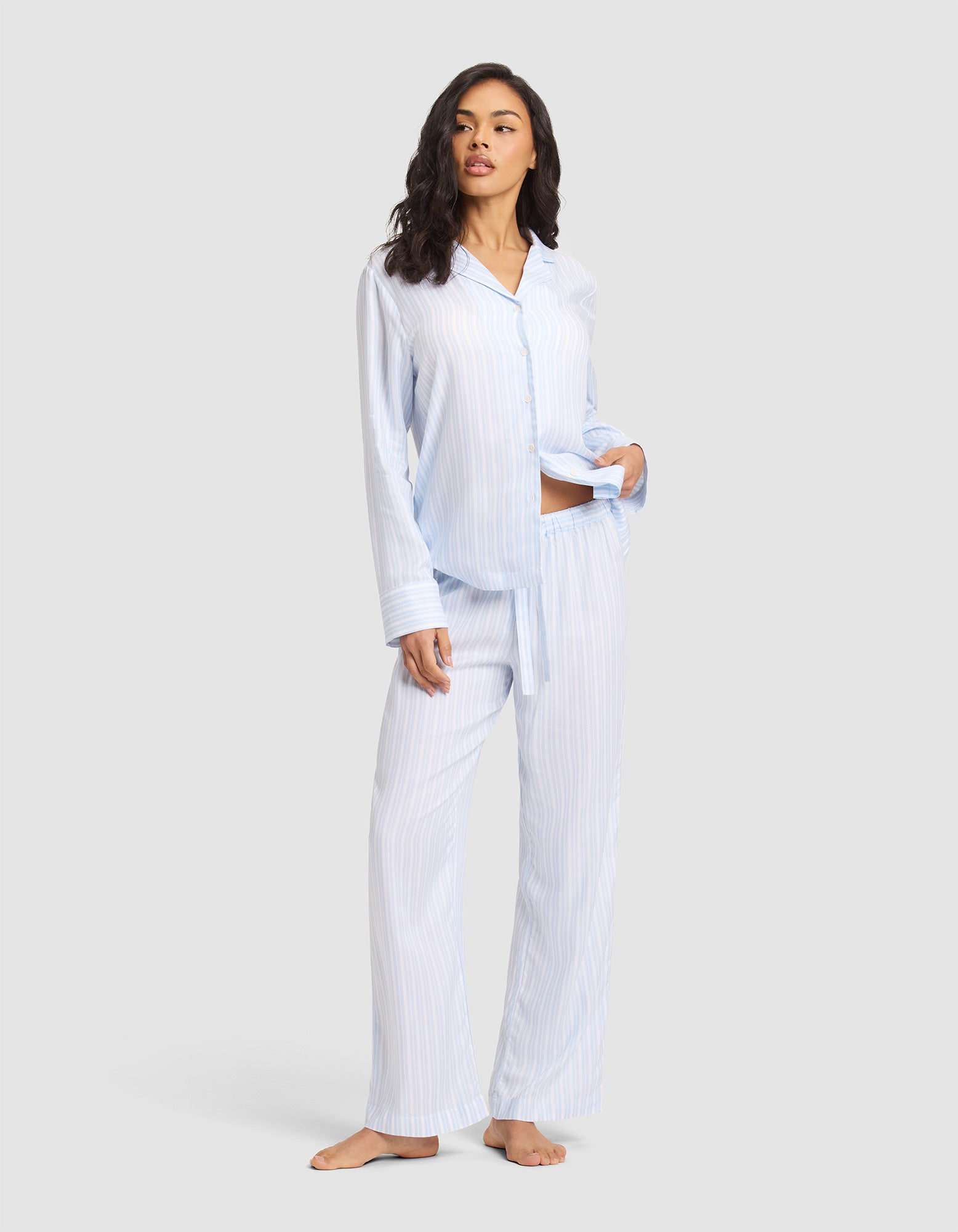 A woman stands barefoot against a light gray background wearing Cozy Earth Women's Soft Woven Pajama Pant in light blue and white stripes with a matching button-up shirt, her left hand in her pocket.  