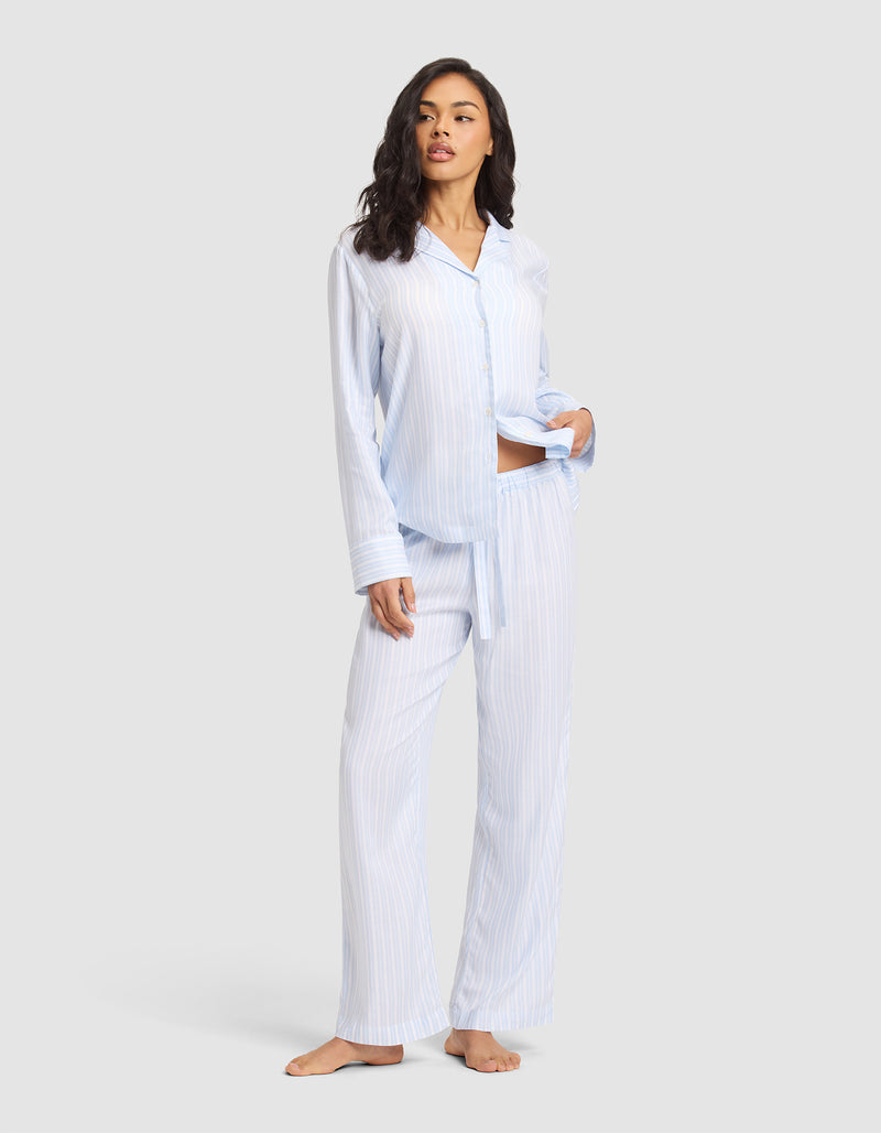 A woman stands barefoot against a light gray background wearing Cozy Earth Women's Soft Woven Pajama Pant in light blue and white stripes with a matching button-up shirt, her left hand in her pocket.
