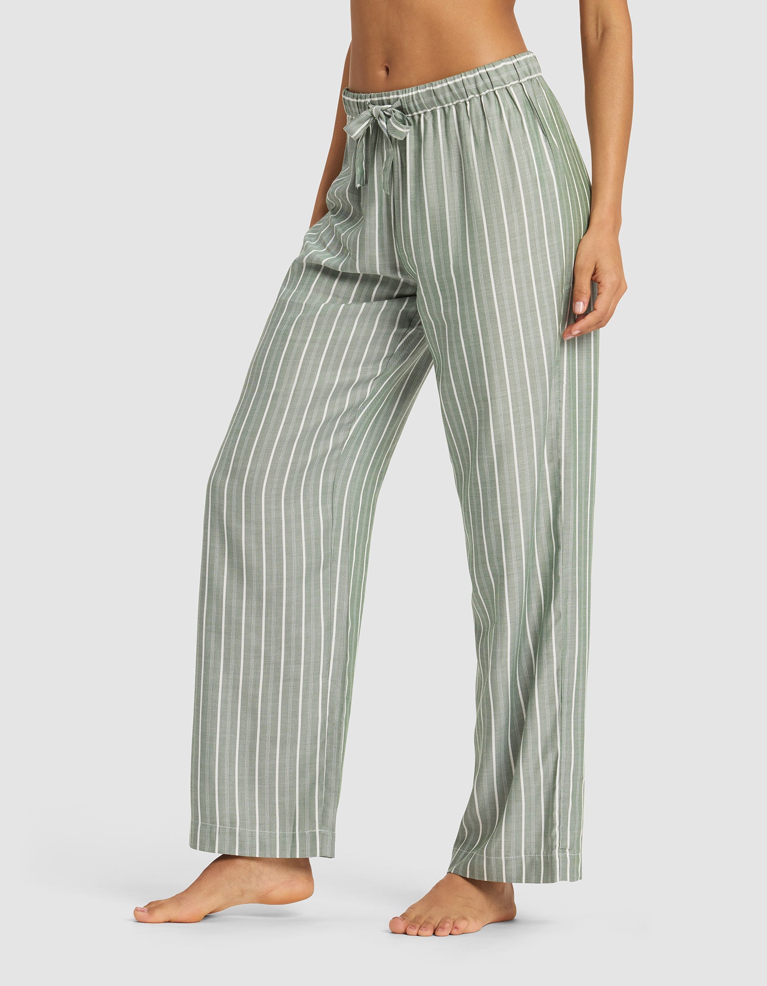 A woman stands barefoot in Cozy Earth's Women's Soft Woven Long Sleeve Pajama Set, featuring loose, light green pants with vertical white stripes and a drawstring waist, against a plain background.