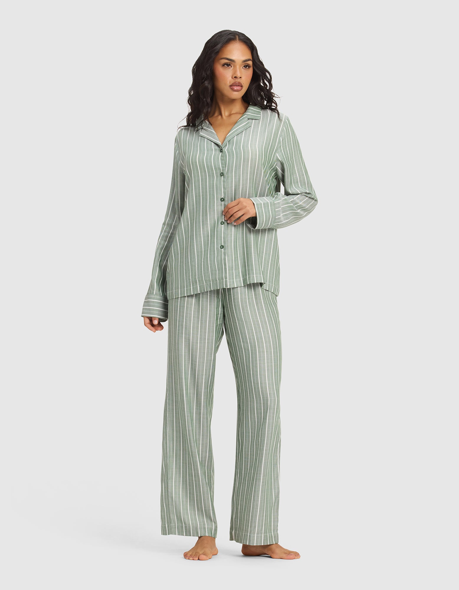 A woman with wavy hair and a neutral expression models Cozy Earth's Women's Soft Woven Pajama Pant, featuring loose-fitting, green and white vertical stripes alongside a matching button-up top, standing barefoot against a plain background.