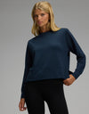 A woman models the Cozy Earth Women’s StudioLite Crewneck in dark blue, paired with black leggings, as she poses with one hand on her hip against a plain gray background.|Color:Eclipse