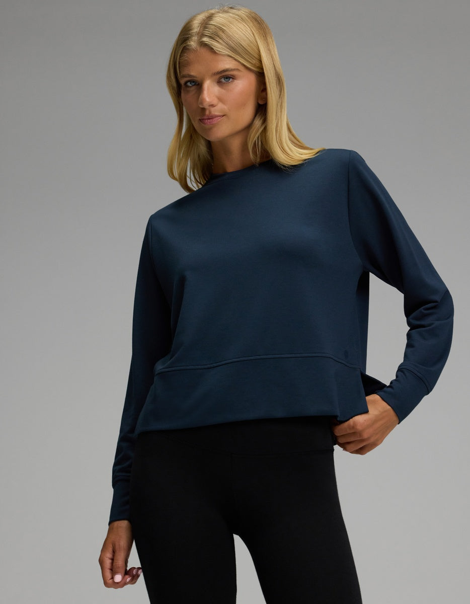 A woman models the Cozy Earth Women’s StudioLite Crewneck in dark blue, paired with black leggings, as she poses with one hand on her hip against a plain gray background.