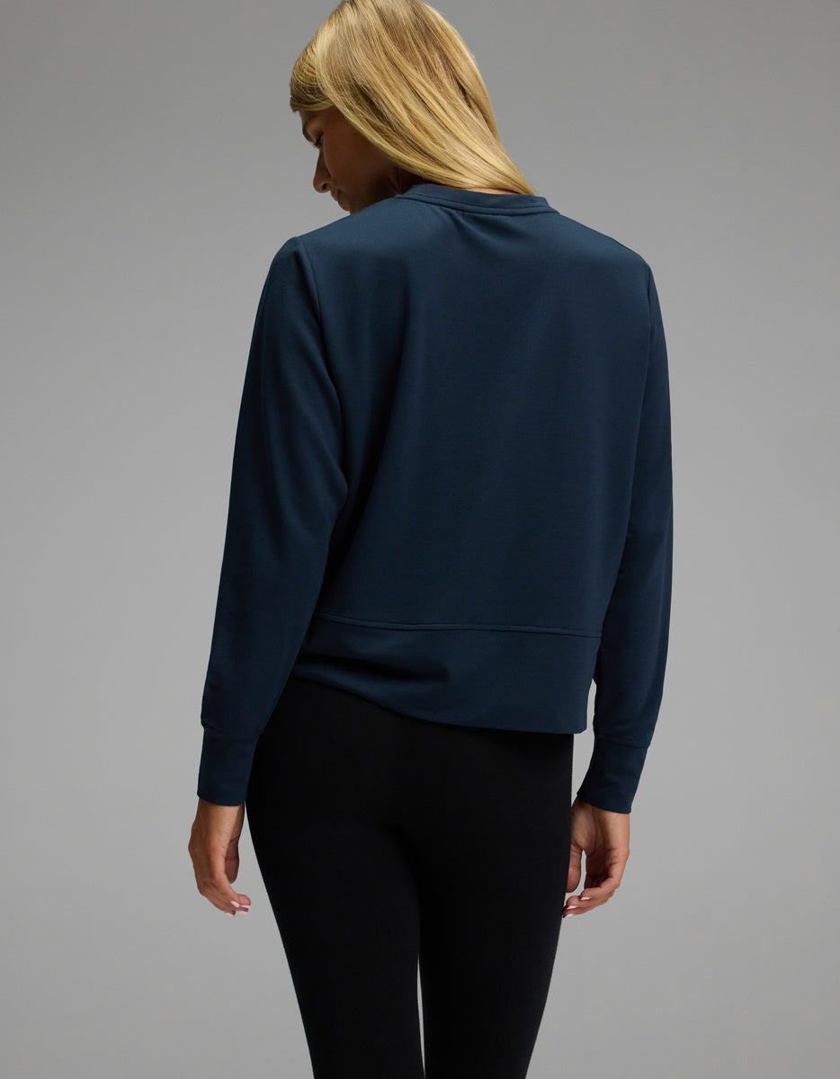 A woman with long blonde hair, seen from behind against a plain gray background, models the Cozy Earth Women’s StudioLite Crewneck in dark blue paired with black leggings.
