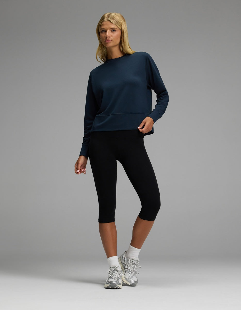 A person stands against a plain gray background, wearing the Cozy Earth Women’s StudioLite Crewneck in navy, paired with black capri leggings, white socks, and gray sneakers. They look slightly to the side with one hand on their hip.
