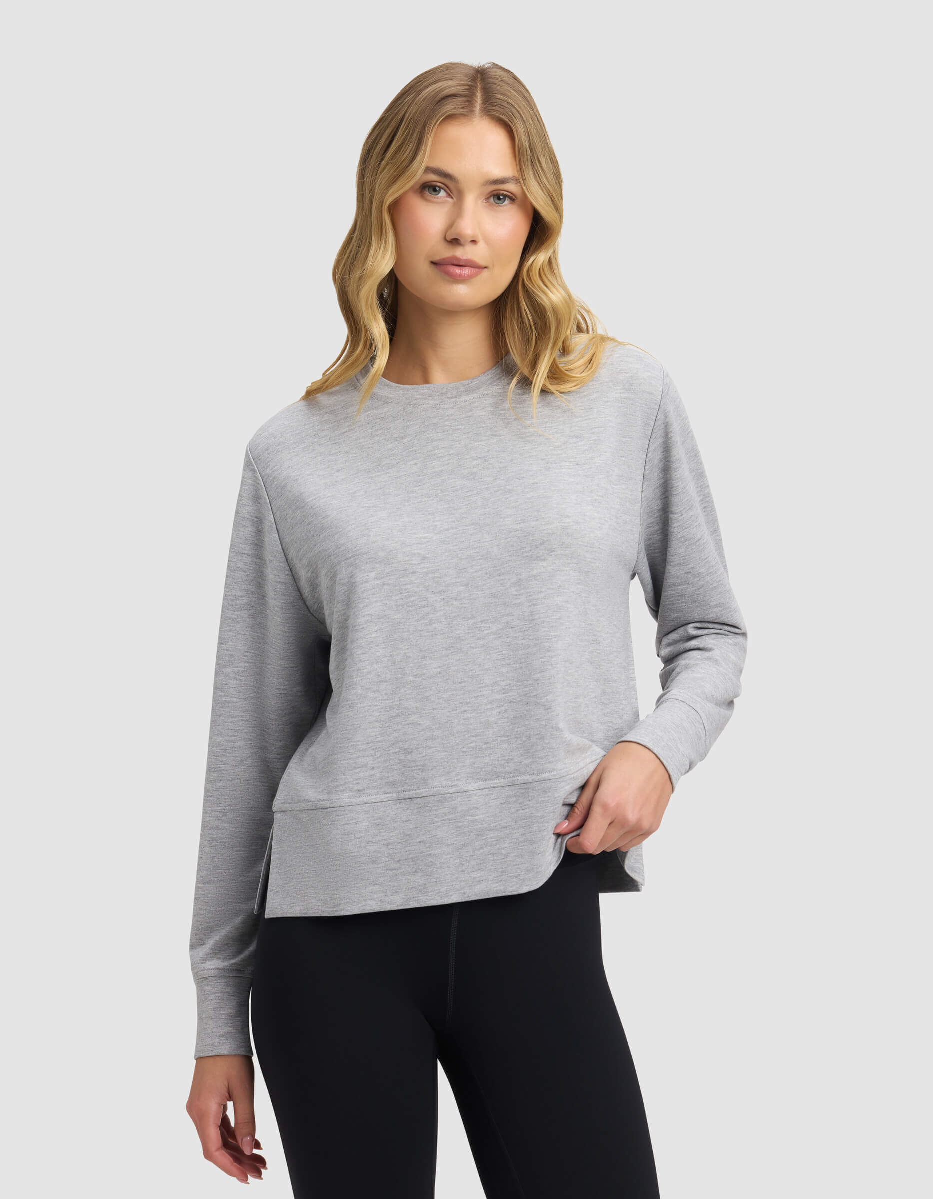 A woman with wavy blonde hair wears the Cozy Earth Women's StudioLite Crewneck in light gray with black leggings, standing against a plain light gray background and displaying a relaxed, neutral expression.|Color:Heather Grey