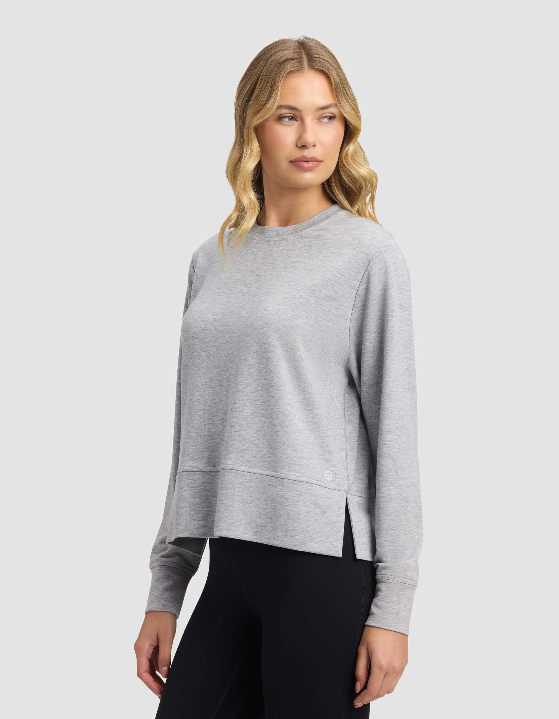 A woman with wavy blonde hair wears the Cozy Earth Women's StudioLite Crewneck in light gray with black leggings, standing against a plain light gray background and looking slightly to her left.|Color:Heather Grey