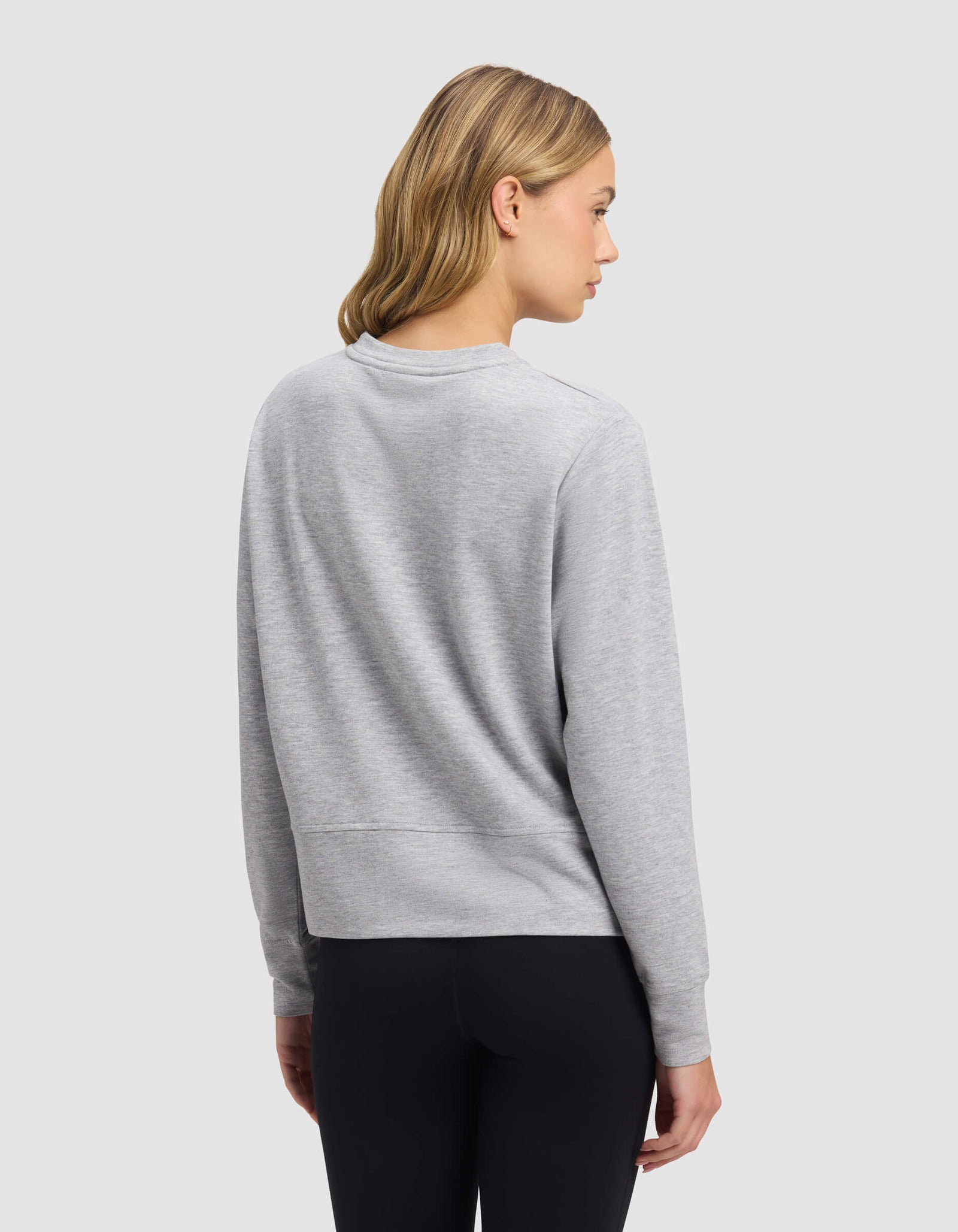 A woman with shoulder-length blonde hair faces away, wearing the Cozy Earth Women's StudioLite Crewneck in light gray with black leggings, set against a plain light gray background.