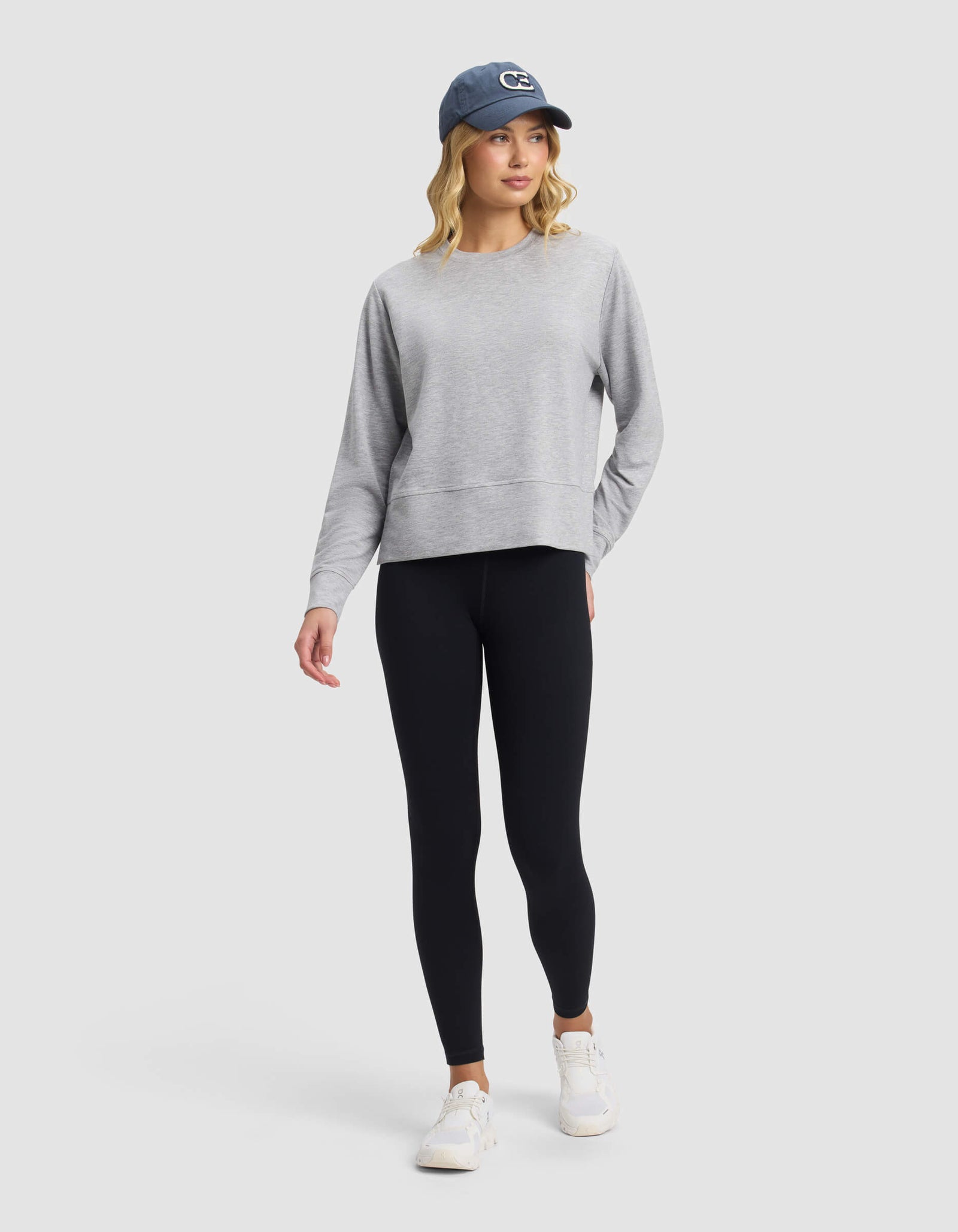 A woman wears the Cozy Earth Women's StudioLite Crewneck, paired with black leggings, white sneakers, and a blue baseball cap. She stands against a light gray background, looking slightly to the side with one hand in her pocket.
