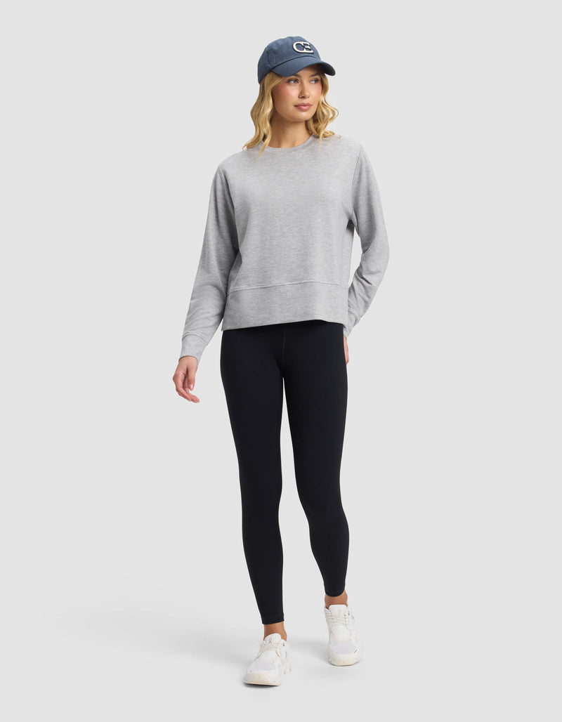 A woman wears the Cozy Earth Women's StudioLite Crewneck, paired with black leggings, white sneakers, and a blue baseball cap. She stands against a light gray background, looking slightly to the side with one hand in her pocket.