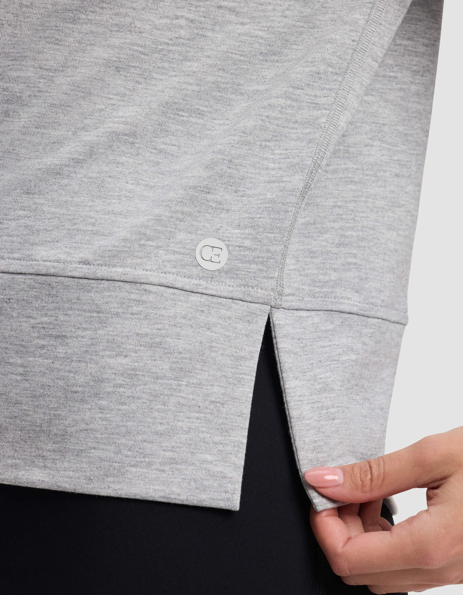 A close-up of someone wearing Cozy Earth's Women's StudioLite Crewneck in light gray, featuring a side slit detail and gently pulling the fabric, paired with black pants.