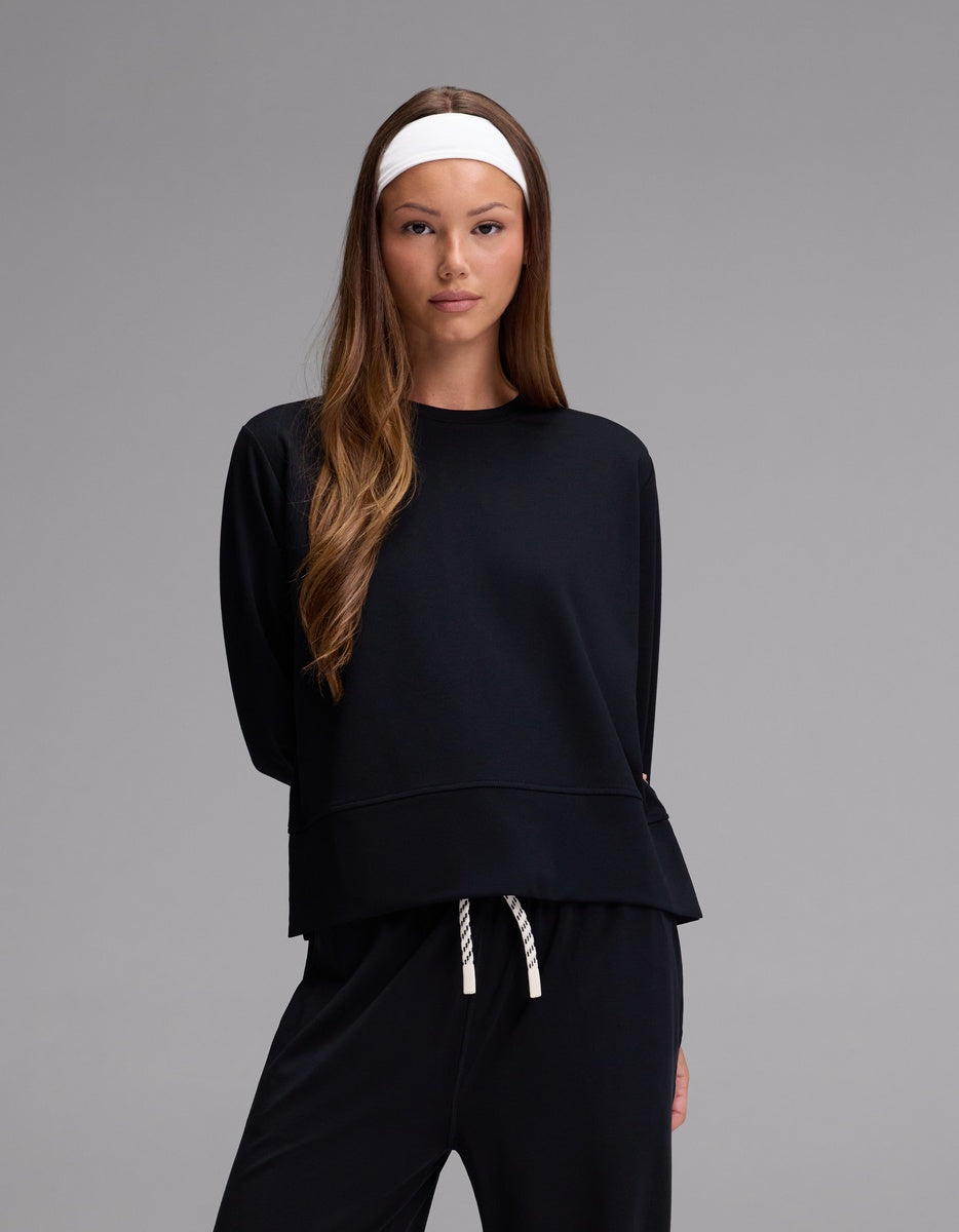 A woman with long brown hair and a white headband wears the Cozy Earth Women’s StudioLite Crewneck in Jet Black, paired with matching pants, standing against a plain gray background.