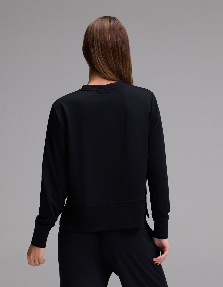 A person with long brown hair, wearing the Cozy Earth Women’s StudioLite Crewneck in black and matching black pants, stands facing away from the camera against a plain gray background.