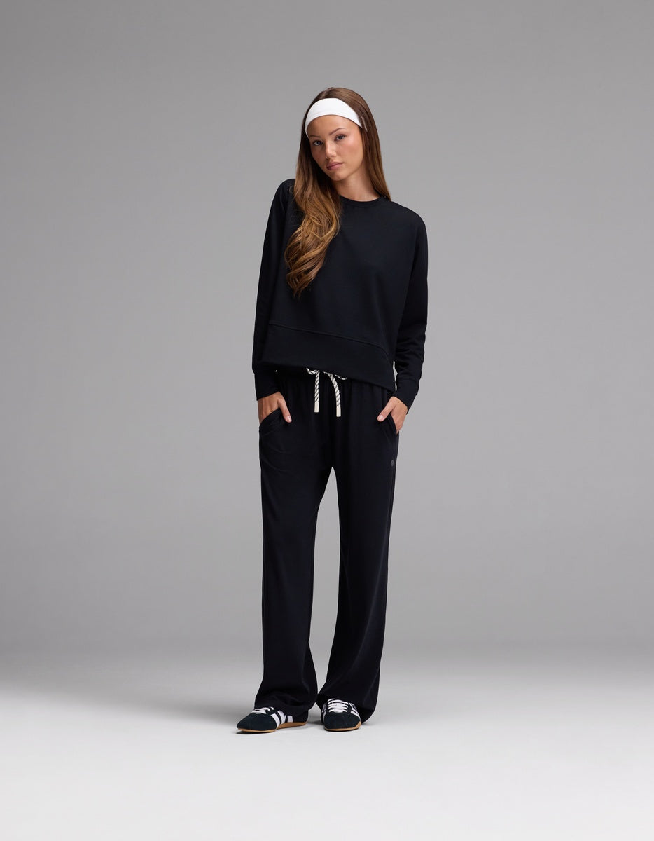 A woman stands against a gray background, wearing the Cozy Earth Women’s StudioLite Crewneck paired with matching black drawstring pants, black sneakers, and a white headband. She has her hands in her pockets and looks straight ahead.
