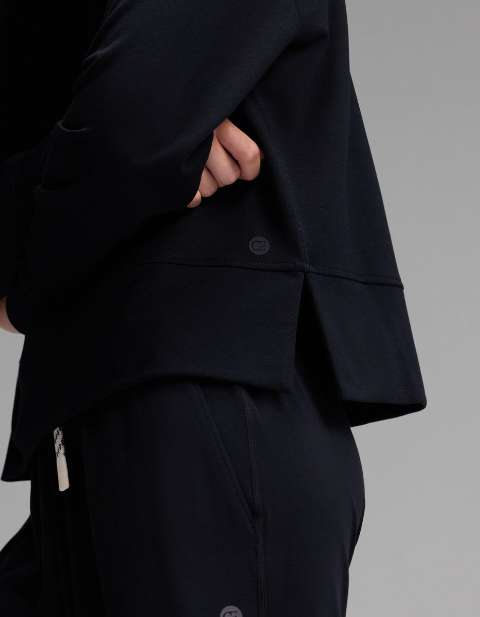 A person wearing the Cozy Earth Women’s StudioLite Crewneck, a loose-fitting black long-sleeve top with a back slit, and matching black pants stands with arms folded against a plain gray background.