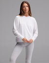 A woman with straight brown hair and a neutral expression stands against a plain gray background, wearing the Cozy Earth Women’s StudioLite Crewneck in white and light gray leggings.|Color:White