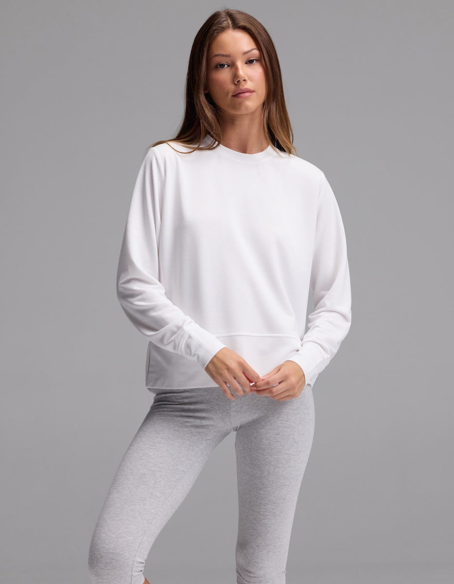 A woman with straight brown hair and a neutral expression stands against a plain gray background, wearing the Cozy Earth Women’s StudioLite Crewneck in white and light gray leggings.