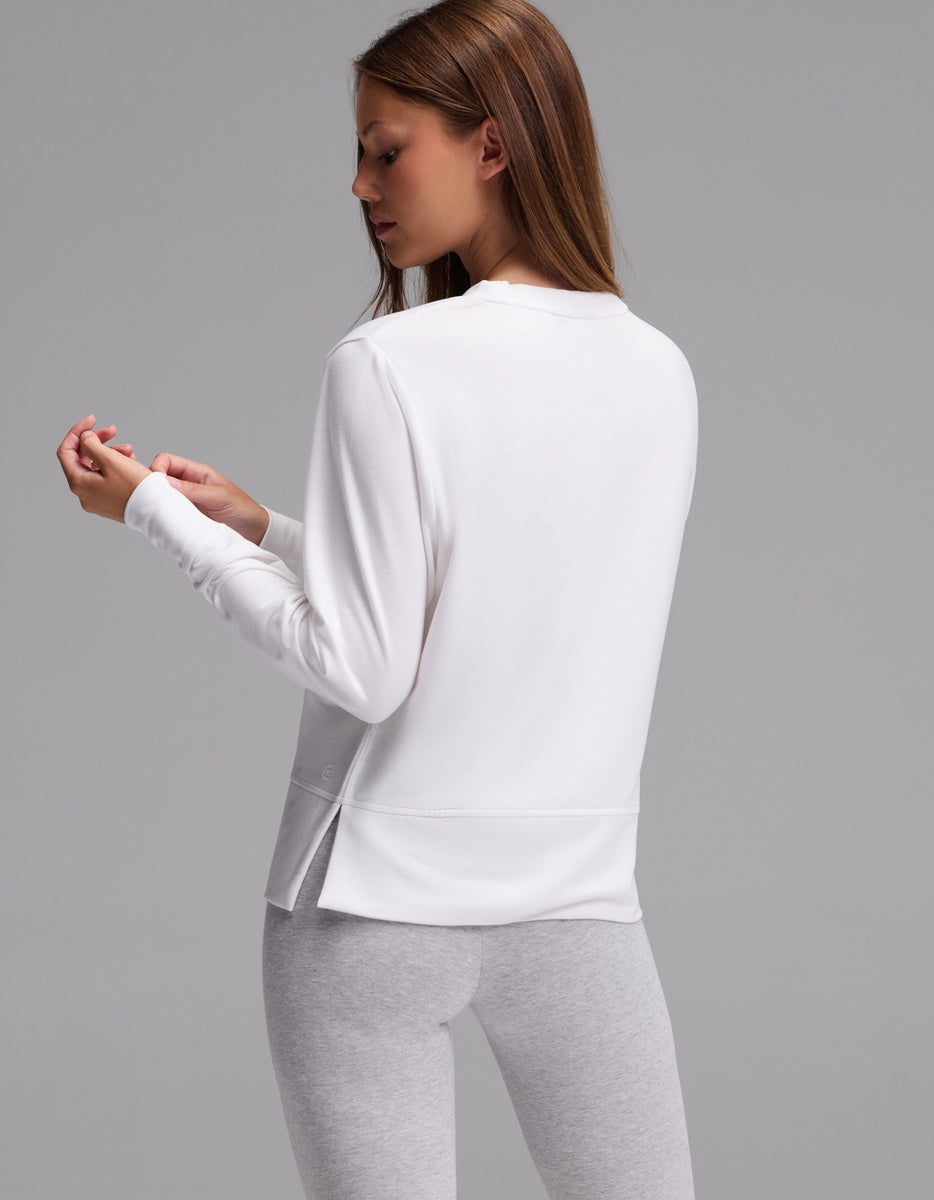 A woman with long brown hair is standing sideways against a plain gray background, wearing the Cozy Earth Women’s StudioLite Crewneck in white and light gray leggings, while looking at her hand.