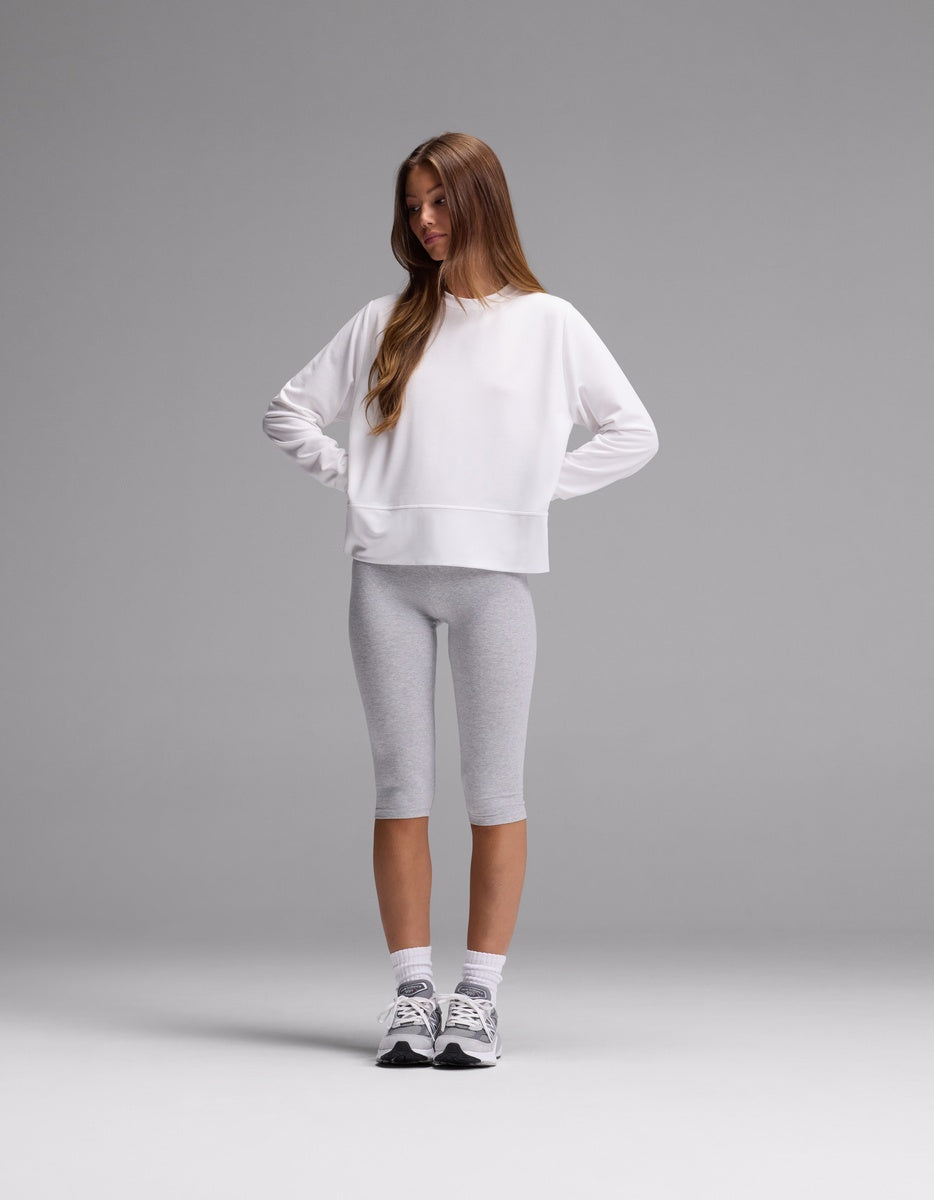 A young woman stands with hands on hips, wearing the Cozy Earth Women’s StudioLite Crewneck in white, paired with light gray knee-length leggings, white socks, and gray sneakers against a plain gray background.