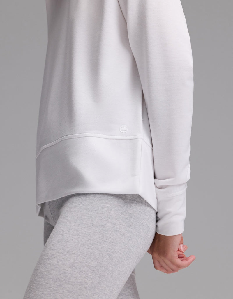 A woman stands sideways against a plain gray background, wearing the Cozy Earth Women’s StudioLite Crewneck in white and light gray leggings, with one arm relaxed at her side.
