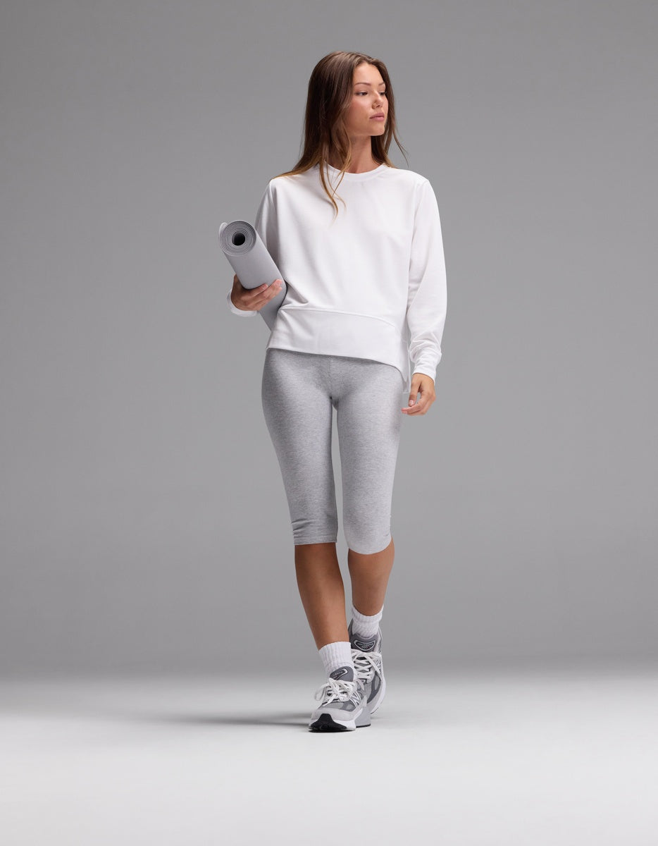 A woman wearing the Cozy Earth Women’s StudioLite Crewneck, paired with gray capri leggings and sneakers, holds a rolled yoga mat while standing against a plain gray background.