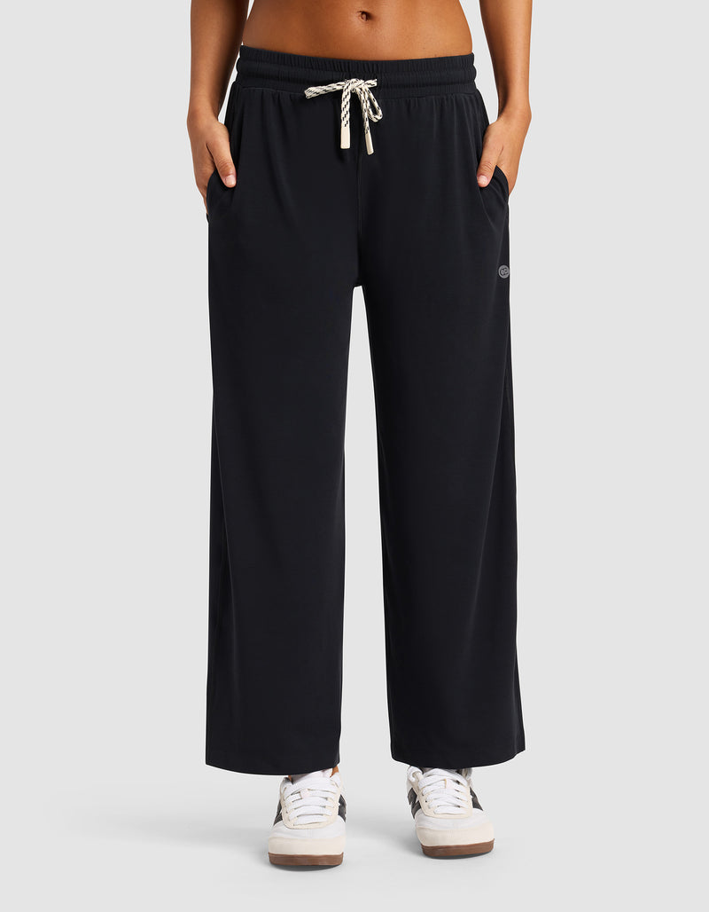 A woman wears Cozy Earth’s Women’s Studio Cropped Wide Leg Pant in black, featuring pockets and a drawstring waist, paired with white sneakers against a plain light gray background.