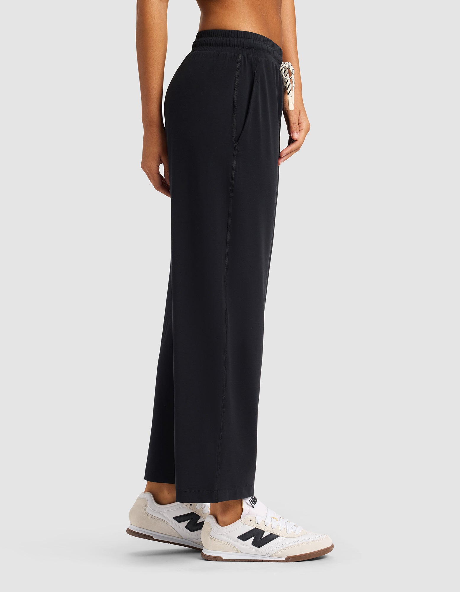 A person wearing Cozy Earth's Women's Studio Cropped Wide Leg Pant in black with white New Balance sneakers, shown from the side against a plain light gray background. Only the lower torso and legs are visible.