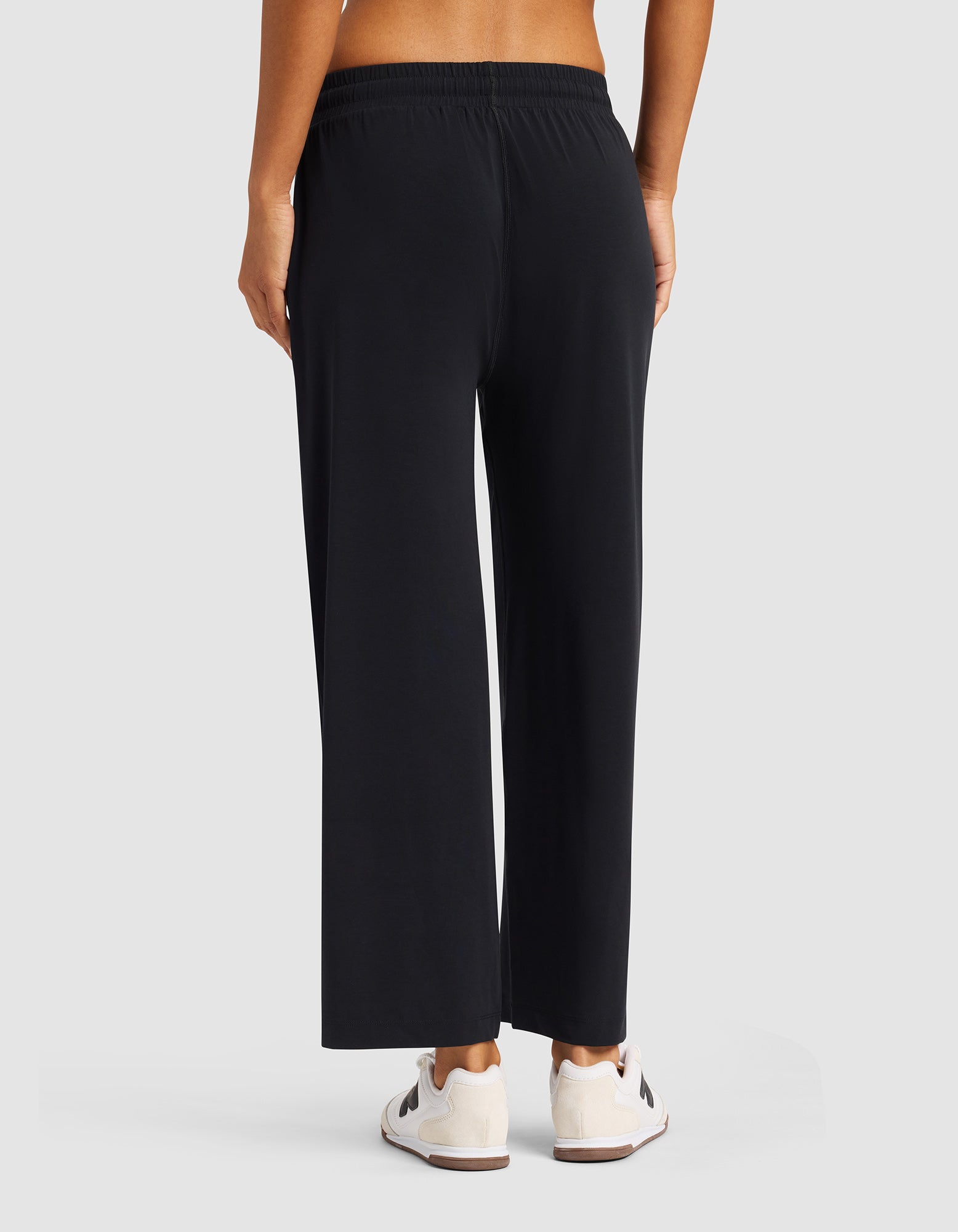 A person stands facing away, wearing Cozy Earth's Women's Studio Cropped Wide Leg Pant in black with an elastic waistband and relaxed fit, paired with white sneakers featuring black accents. The background is plain and light-colored. 