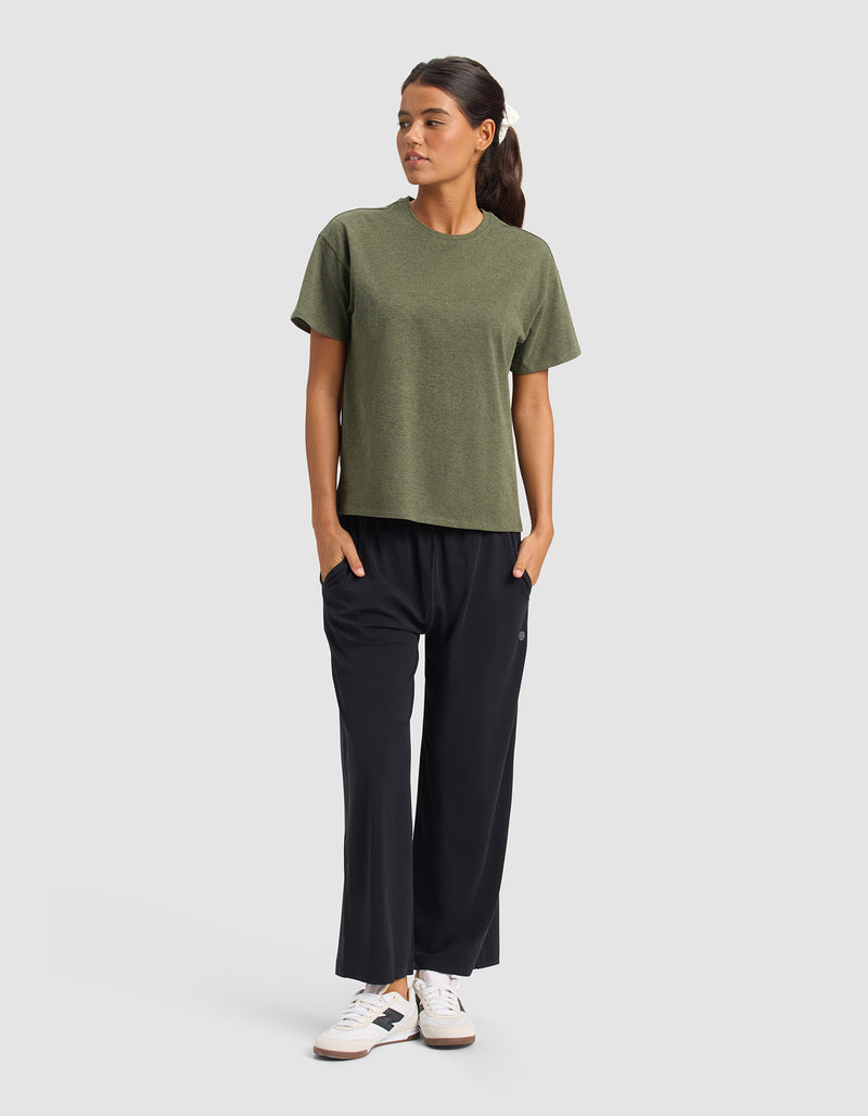 A woman stands against a plain background wearing Cozy Earth’s Women’s Studio Cropped Wide Leg Pant in black, paired with an olive green t-shirt and white sneakers with black accents. Her hands are in her pockets and she gazes to the side neutrally.