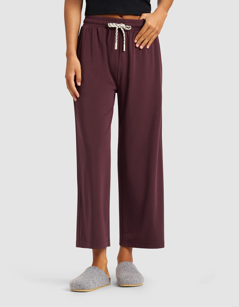 A person wearing Cozy Earth’s Women's Studio Cropped Wide Leg Pant in maroon, paired with a black top and gray slippers, stands against a plain light background.