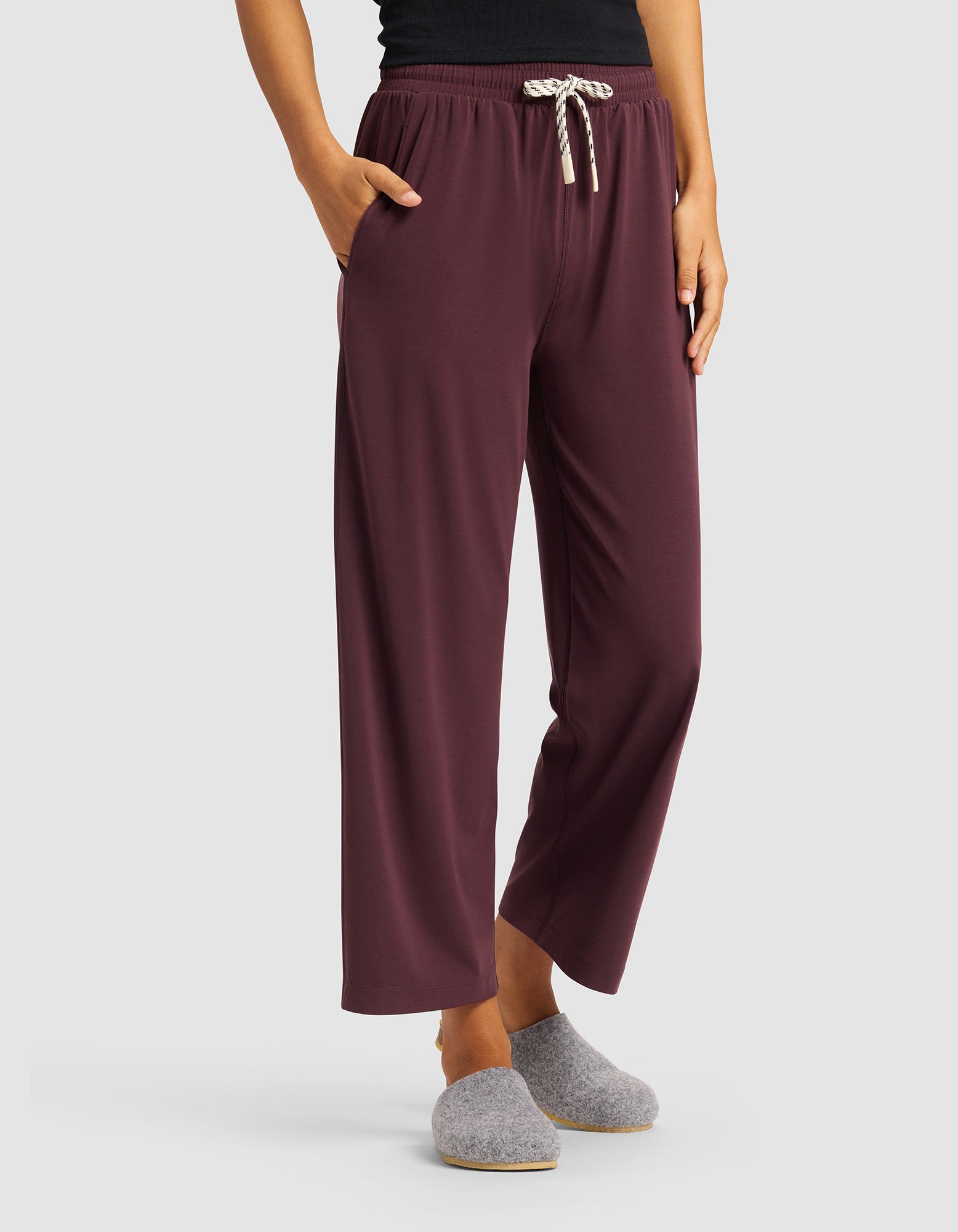 A woman models the Cozy Earth Women's Studio Cropped Wide Leg Pant in maroon with a white drawstring, paired with a black top and gray slippers, standing hands in pockets against a plain light background.