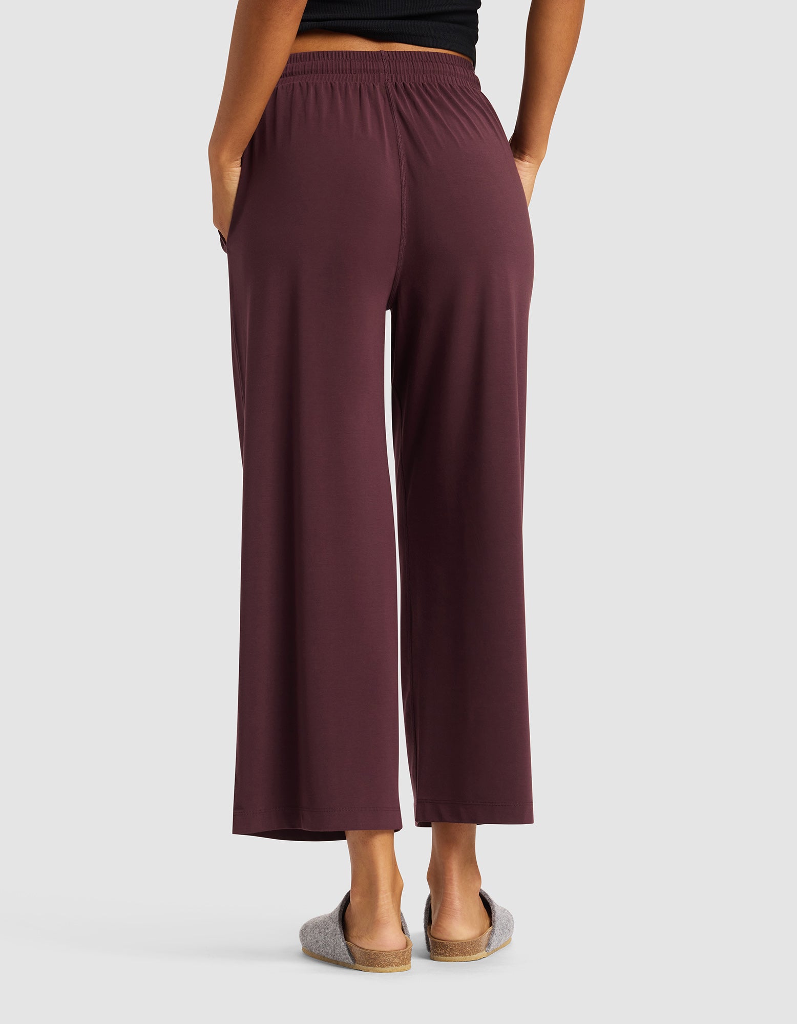 A person stands with their back to the camera, hands in pockets, wearing Cozy Earth's Women's Studio Cropped Wide Leg Pant in maroon, a black top, and gray slippers against a plain light gray background.