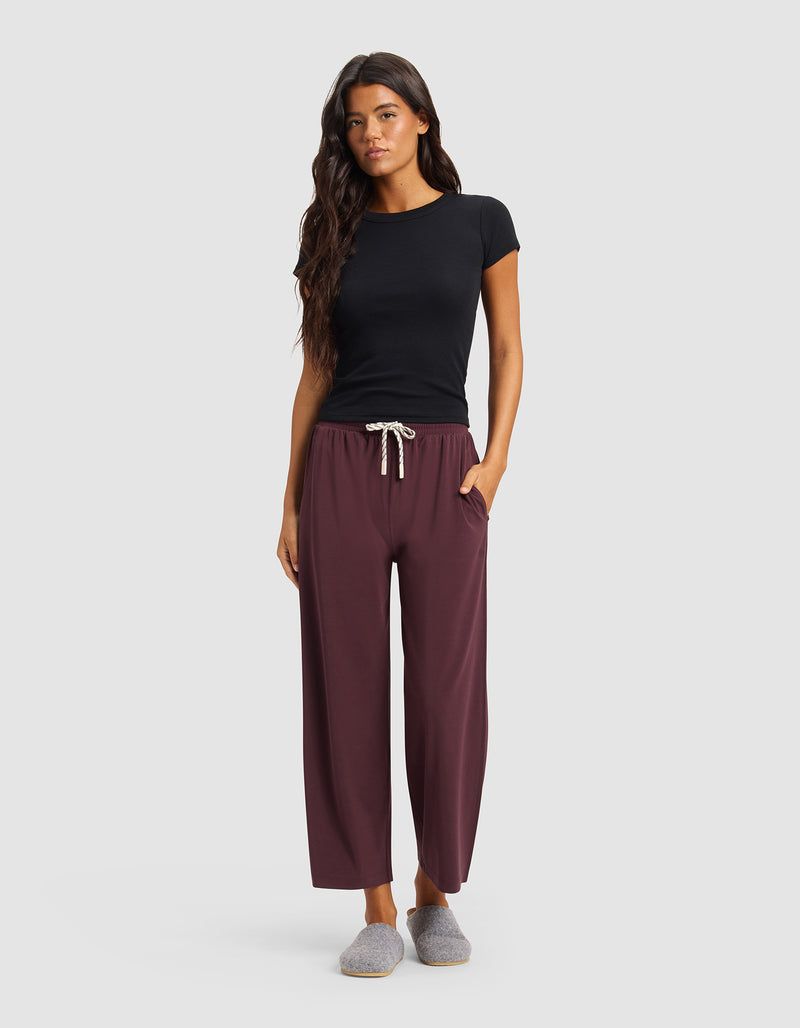 A woman faces forward against a plain light background, wearing Cozy Earth Women's Studio Cropped Wide Leg Pants in burgundy, a fitted black t-shirt, and gray slippers, with one hand in her pocket.