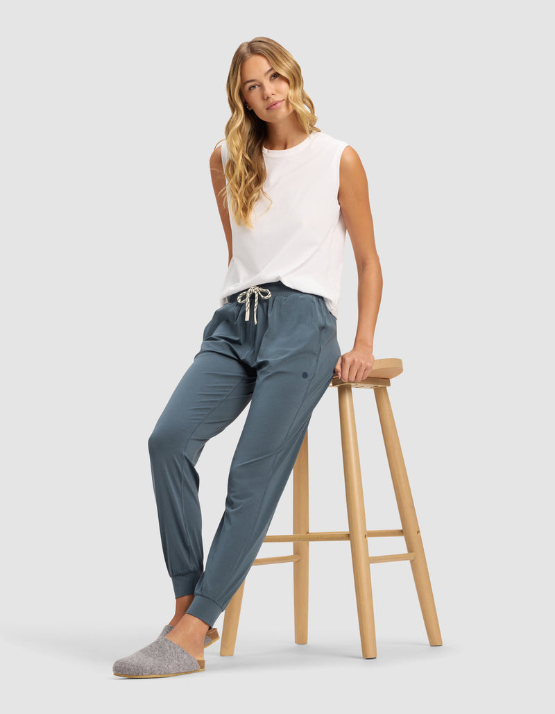 A woman with long blonde hair models Cozy Earth's Women's Studio Jogger in blue, paired with a white sleeveless top and gray slippers, sitting on a wooden stool against a plain light gray background.