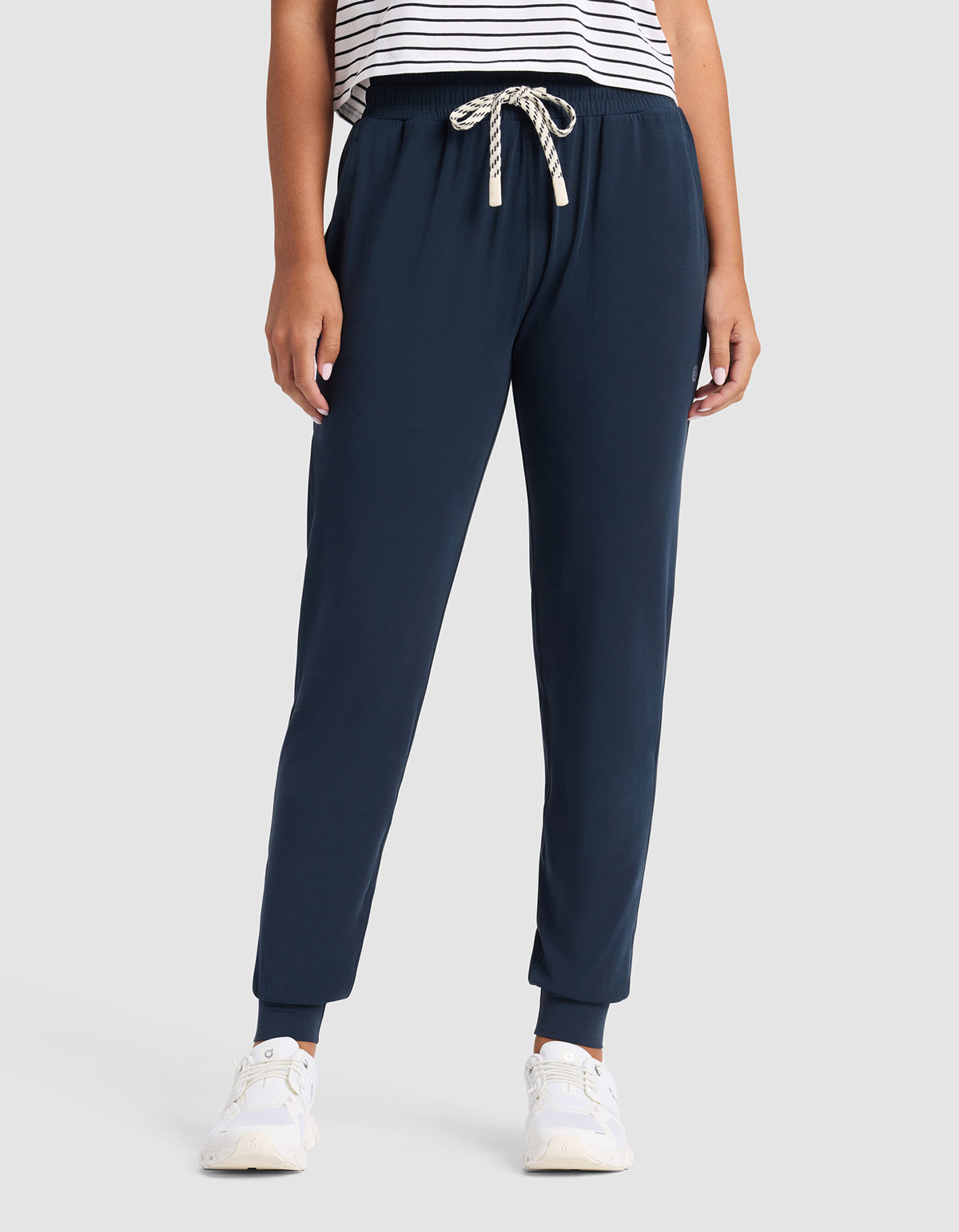 A person models Cozy Earth Women's Studio Jogger in navy blue with a white drawstring, paired with a white and black striped shirt and white sneakers, against a plain light gray background. The individual's head is not visible.|Color:Eclipse