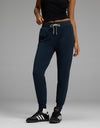 Wearing the Cozy Earth Women’s Studio Jogger in navy, a person stands against a plain gray background, styled with a black sleeveless top, white socks, and black Adidas sneakers.|Color:Eclipse