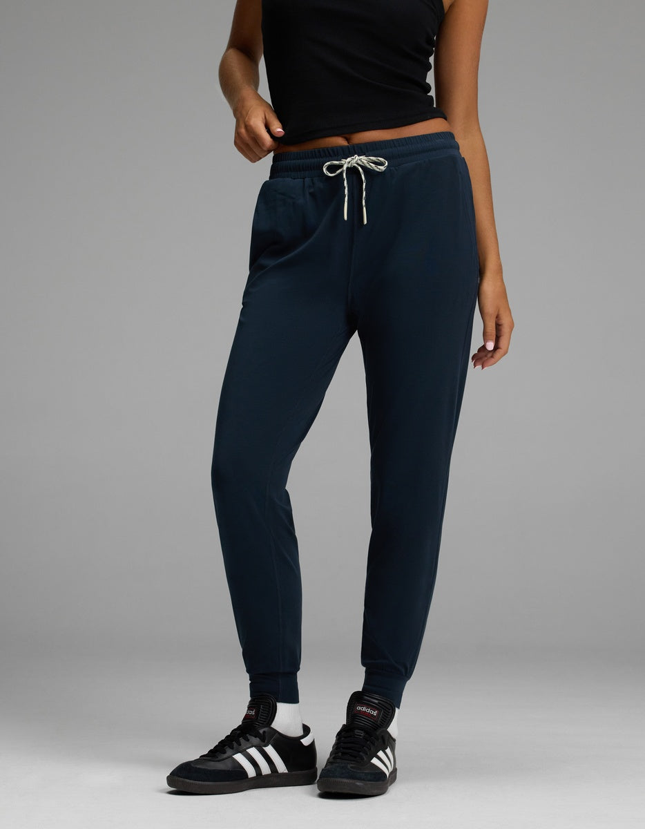 Wearing the Cozy Earth Women’s Studio Jogger in navy, a person stands against a plain gray background, styled with a black sleeveless top, white socks, and black Adidas sneakers.|Color:Eclipse
