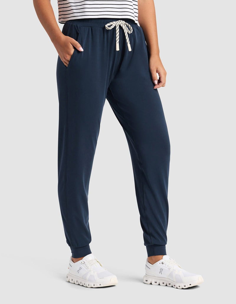 A person wears Cozy Earth's Women's Studio Jogger in navy blue with a drawstring waistband, paired with white athletic shoes and a white top featuring black stripes. Hands are in pockets; only the lower half is visible.