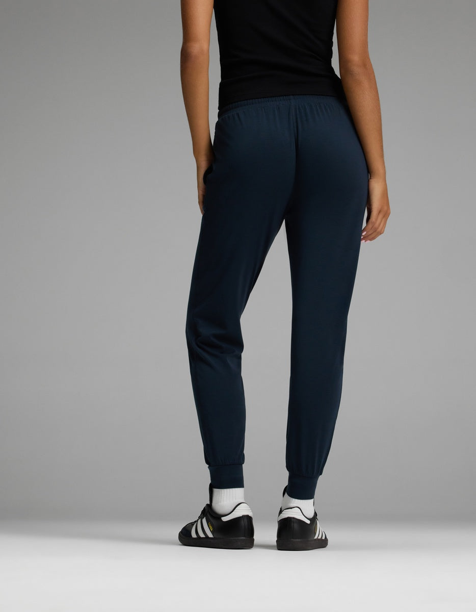 A person wearing a black top, Cozy Earth Women’s Studio Jogger pants in dark blue, and black-and-white athletic shoes stands facing away from the camera against a plain gray background.