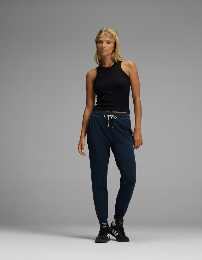 A woman stands against a plain gray background, wearing Cozy Earth’s Women’s Studio Jogger in navy with a drawstring waist, paired with a black sleeveless top and black sneakers.