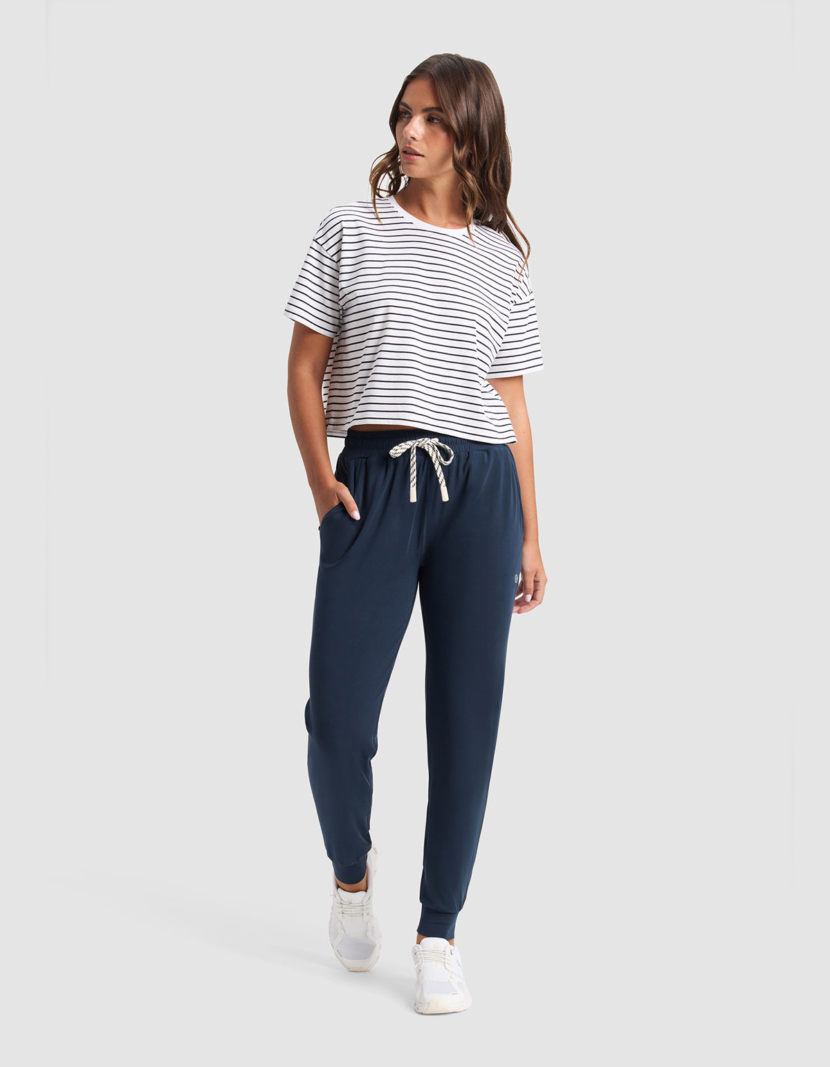 A woman with wavy brown hair stands against a plain background wearing Cozy Earth’s Women's Studio Jogger in navy blue, a white and black striped t-shirt, and white sneakers. Her left hand is in her pocket.|Color:Eclipse