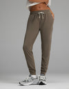 A person is wearing Cozy Earth’s Women’s Studio Jogger in taupe featuring a drawstring waistband, paired with gray sneakers and a white cropped top. The image has a plain gray background and highlights the lower body and legs.|Color:Granite