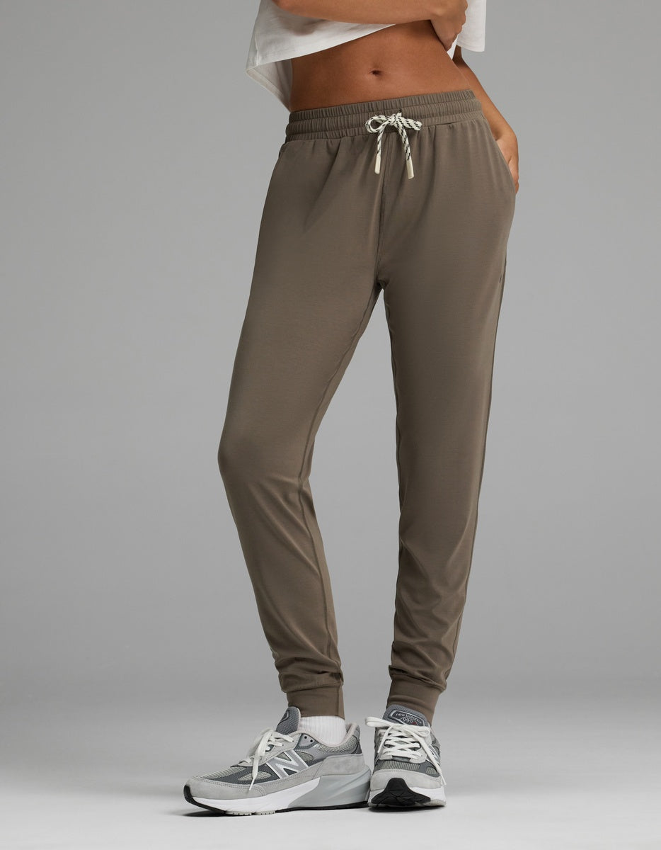A person is wearing Cozy Earth’s Women’s Studio Jogger in taupe featuring a drawstring waistband, paired with gray sneakers and a white cropped top. The image has a plain gray background and highlights the lower body and legs.|Color:Granite
