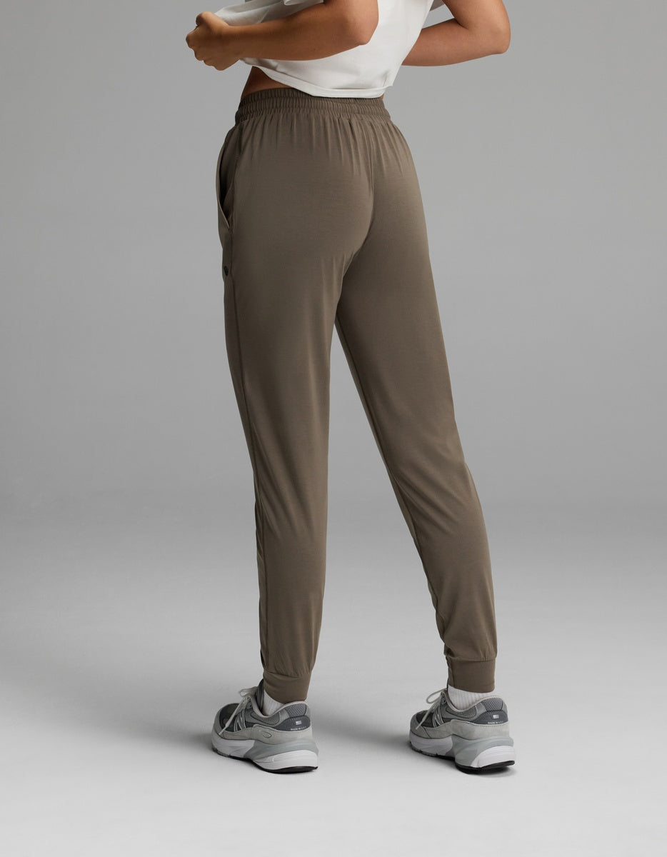 A person is seen from behind wearing the Cozy Earth Women’s Studio Jogger in taupe, paired with gray sneakers and a white top. The image highlights the fit and style of the pants against a plain gray background.|Color:Granite