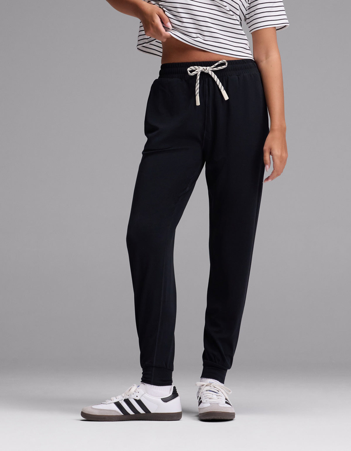 A person wearing a striped short-sleeve shirt, white sneakers with black stripes, and the Cozy Earth Women’s Studio Jogger in Jet Black stands against a plain gray background.