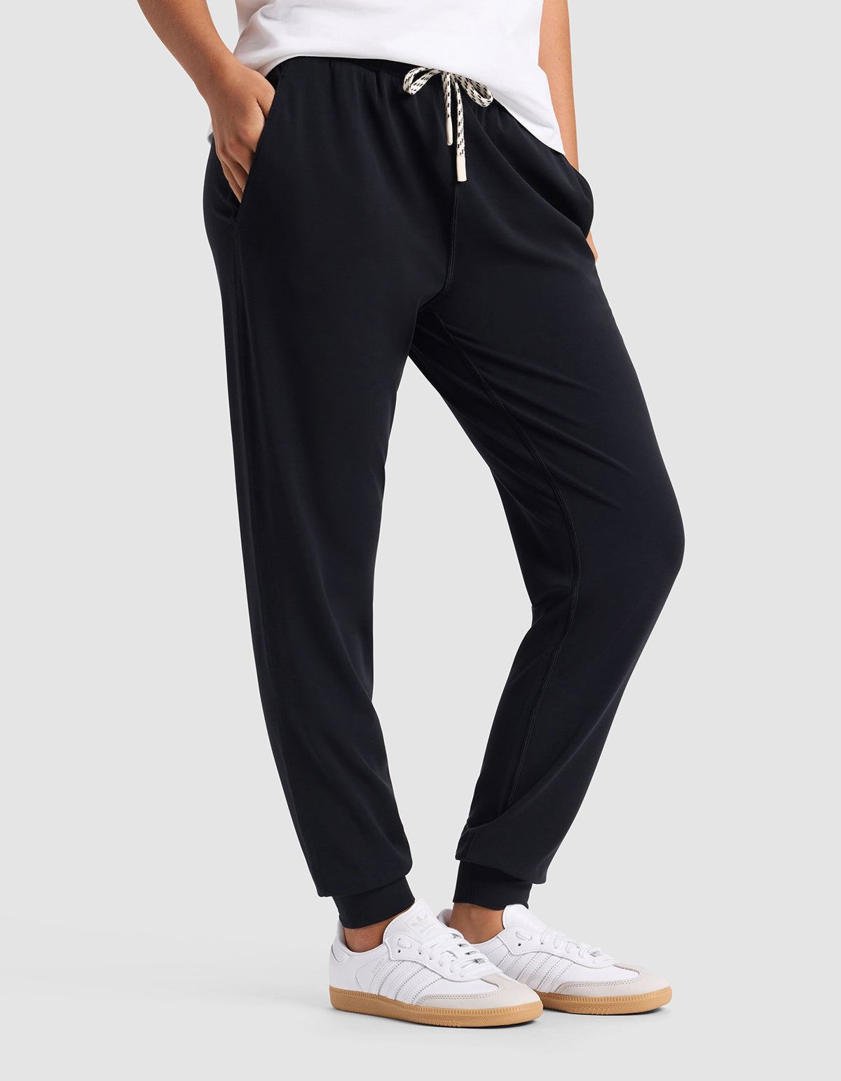 A woman wears Cozy Earth Women's Studio Jogger—black with a drawstring waist—paired with white sneakers and a white top, standing hands-in-pockets against a plain light gray background.|Color:Jet Black